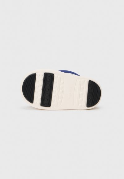 Fehér sneaker talp fekete gumi tapadóbetétekkel. Ki van emelve egy logó, amelyet sima, lekerekített fehér szegély vesz körül.