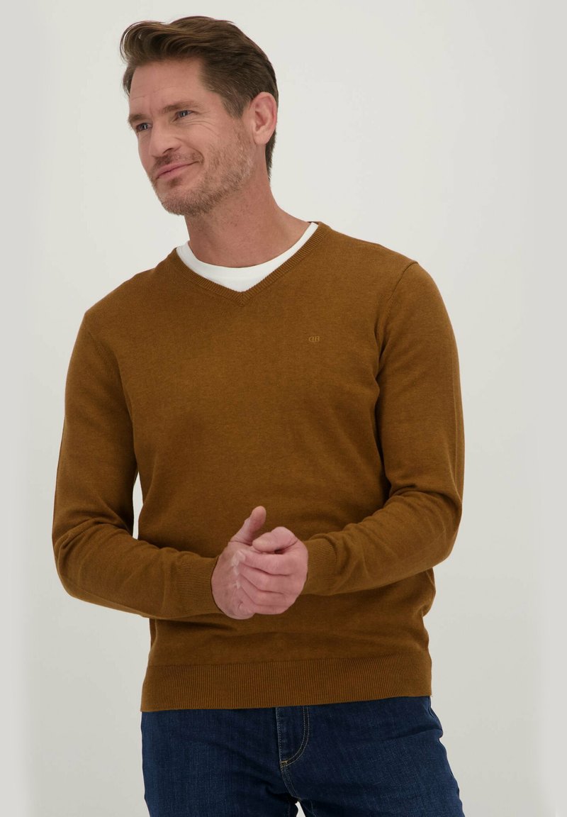 Bruine, gebreide trui met een V-halsontwerp en geribbelde manchetten, gedragen over een witte shirt, gecombineerd met blauwe spijkerbroek. Eenvoudige, naadloze textuur.