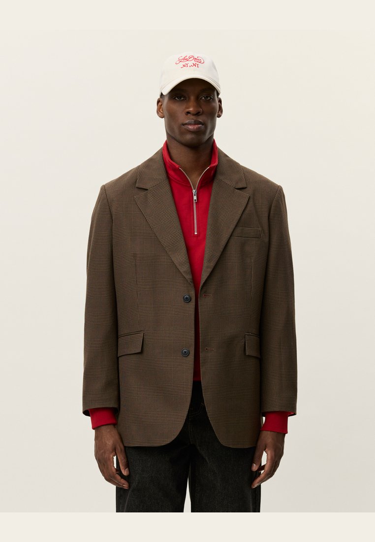 Brunternet blazer med notch-lapel og to knapper, båret over en rød zip-up top. Bomulds cap med broderet hvidt logo.