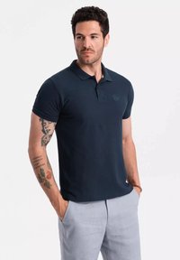 Polo de color navy con mangas cortas, cuello plano y un sutil detalle de logo. La tela parece suave y ajustada, combinada con pantalones claros.