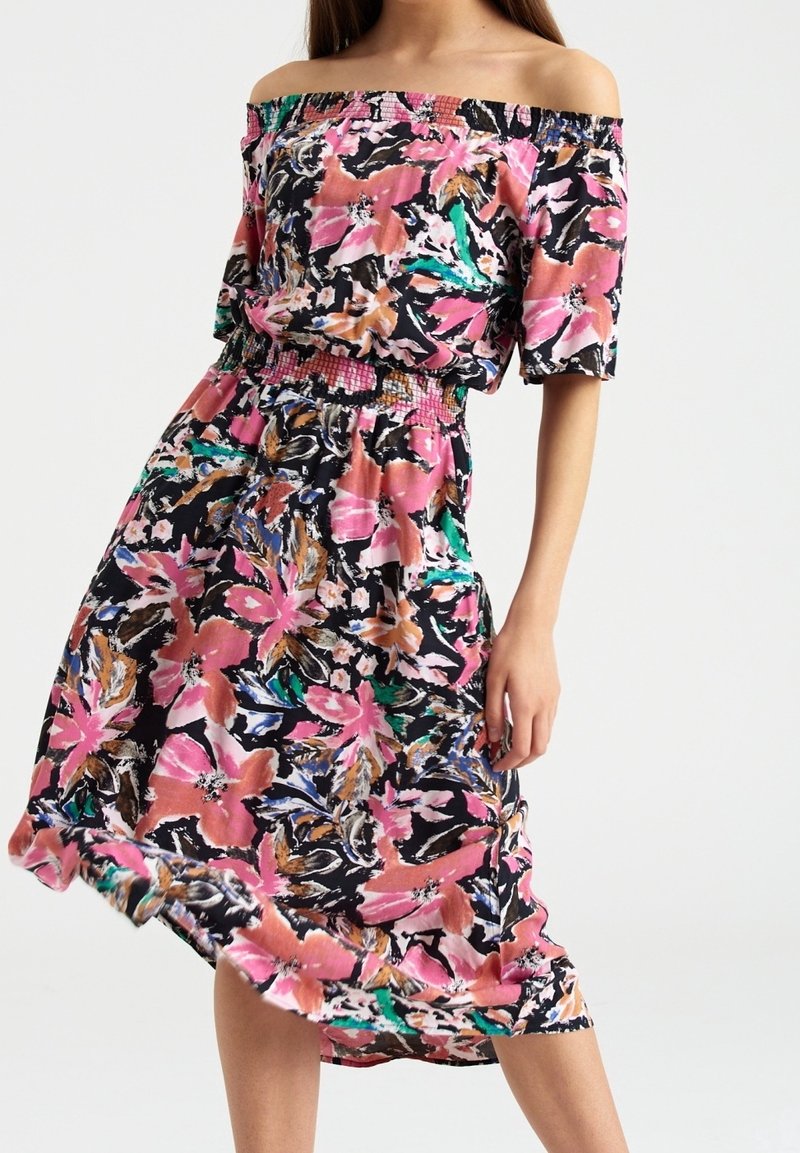 Femme portant une robe fleurie à épaules dénudées avec des motifs roses, noirs, verts et oranges, se tenant devant un fond uni.