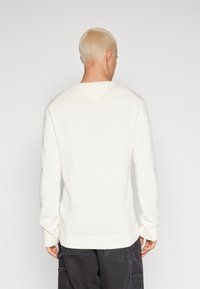 Jersey crema con mangas largas, puños y dobladillo acanalados, que presenta un detalle de yugo en forma de V en la parte posterior. Textura de punto suave, corte informal.