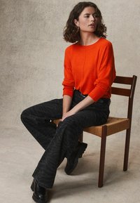 Orangener Strickpullover mit langen Ärmeln, kombiniert mit schwarzen Jeans im Zebra-Muster mit weitem Bein und schwarzen Stiefeletten, sitzt auf einem Holzstuhl.