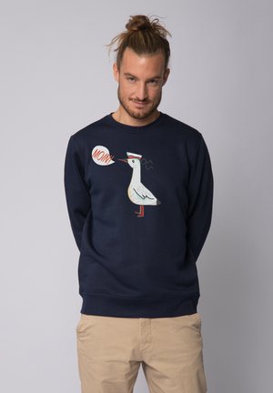 Navyblauwe sweatshirt met een witte zeevogelafbeelding met rode accenten en een spreekwolk met de tekst "MONN!" Zachte stof, ronde halsontwerp.
