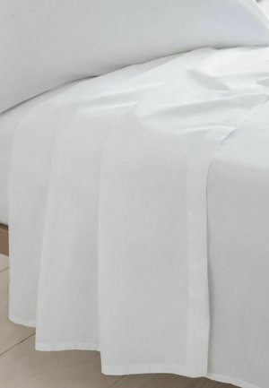 Weißes Bettlaken, das über den Rand eines hölzernen Bettrahmens drapiert ist, mit einem teilweise sichtbaren weißen Kissen auf einem hellen Boden.