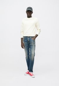 Μάλλινο πουλόβερ σε κρεμ απόχρωση, ξεβαμμένο μπλε τζιν και κόκκινα αθλητικά παπούτσια. Το σύνολο διαθέτει καθημερινό σχέδιο με στρογγυλή λαιμόκοψη και χαλαρή εφαρμογή.