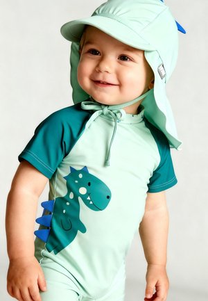 Kleinkind lächelt, trägt ein hellgrünes, dinosaurusmotiviertes Badeoutfit mit blauen Stacheln und einen breitkrempigen Hut, der unter dem Kinn gebunden ist.