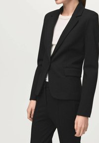 Kobieta w dopasowanej czarnej marynarce i pasujących czarnych spodniach, na białym tle, stojąca na jasnym, gładkim tle.