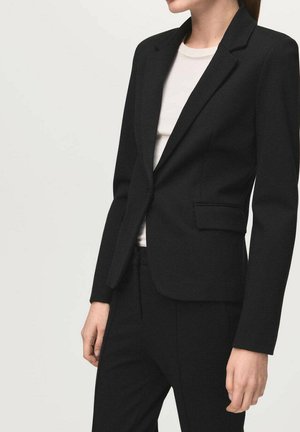 Kobieta w dopasowanej czarnej marynarce i pasujących czarnych spodniach, na białym tle, stojąca na jasnym, gładkim tle.