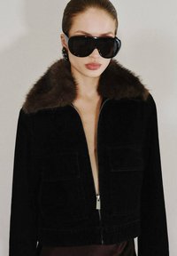 Černá kordurová bunda s hnědým koženým límcem, zipovým zapínáním a dvěma předními kapsami. Nositelná na odhaleném hrudníku, doplněná velkými černými slunečními brýlemi.