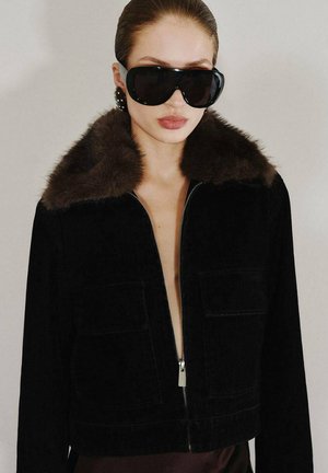 Veste en velours côtelé noir avec un col en fourrure marron, fermeture éclair et deux poches avant. Portée sur un torse nu, accompagnée de grandes lunettes de soleil noires.