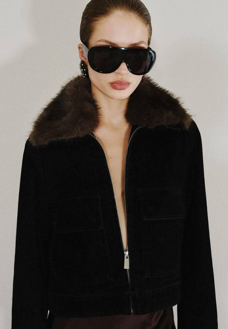 Černá kordurová bunda s hnědým koženým límcem, zipovým zapínáním a dvěma předními kapsami. Nositelná na odhaleném hrudníku, doplněná velkými černými slunečními brýlemi.