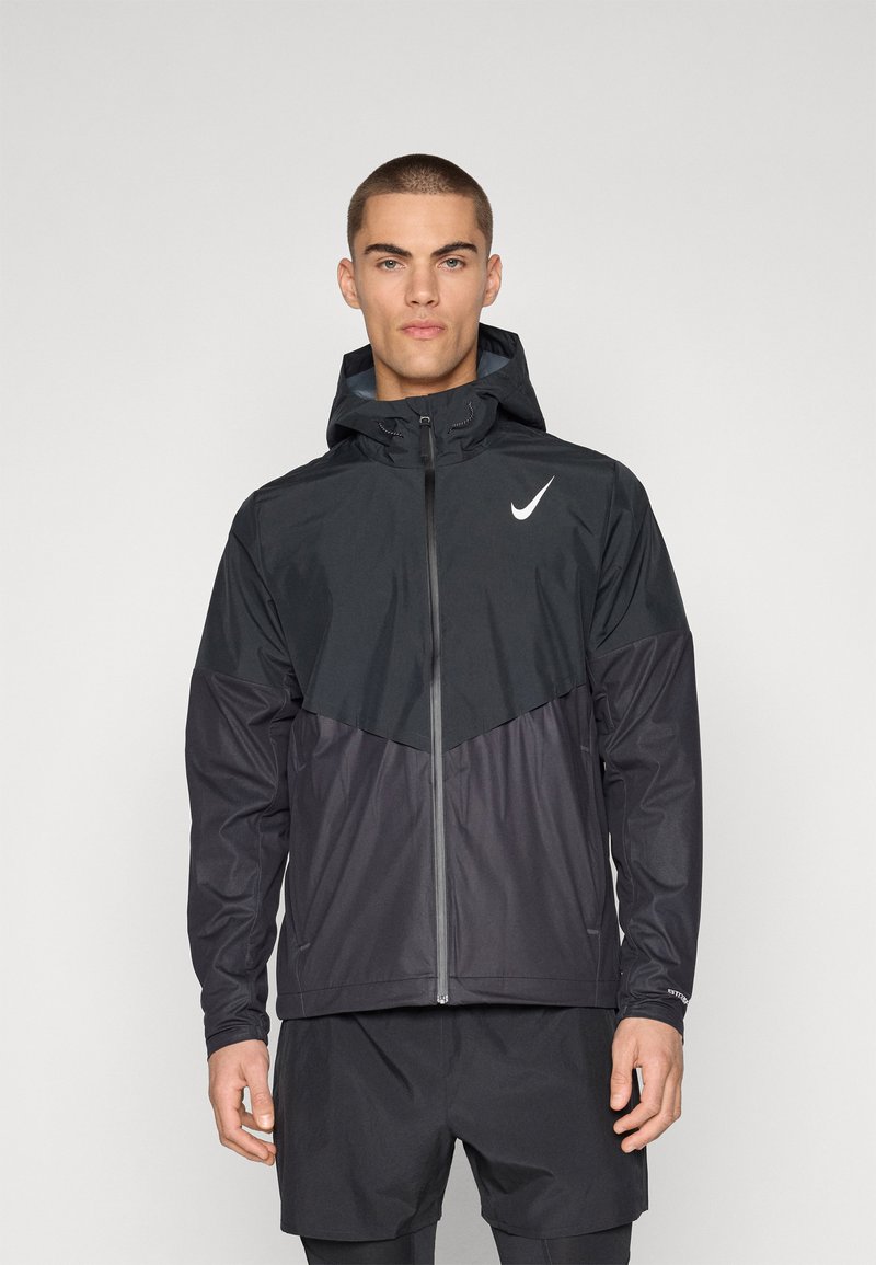 Giacca a vento con cappuccio Nike nera con un motivo triangolare grigio sul petto. Tessuto morbido, zip intera, polsini elasticizzati e cappuccio regolabile.