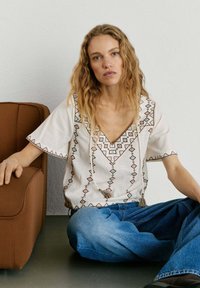 Mladá žena s vlnitými vlasmi sedí na podlahe v tureckom sede, oblečená v bielej vyšívanej blúzke a modrých džínsoch, opierajúc ruky o hnedé kreslo.