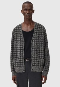 Grå og svart rutete cardigan laget av lodden materiale, med V-hals, frontlommer, og knappeåpning. Brukt over en svart skjorte.