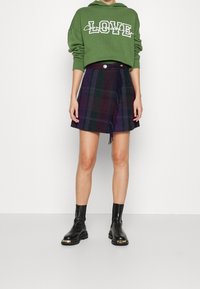 Groene capuchontrui met witte "LOVE" tekst, gecombineerd met een geruite minirok in paars en groen, met franjes, en zwarte enkellaarsjes.