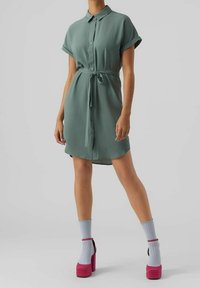 Vihreä lyhythihainen paitamekko, jossa on kaulus ja solmittu vyötärö. Mekossa on napillinen etuosa ja laskeutuva kangas. Asu täydentyy vaaleanpunaisilla korokepohjakengillä ja sinisillä sukilla.