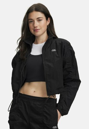 Junge Frau mit langen dunklen Haaren, die eine schwarze kurze Jacke und Hose mit Under Armour Logos sowie ein schwarz-weißes mehrlagiges Top trägt.