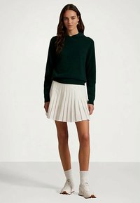 Naine kannab tumerohelist kapuutsiga särki, valgest lohutusseelikust, valgetest sokkidest ja valgestest jalanõudest, seistes ühele värvilisele taustale.