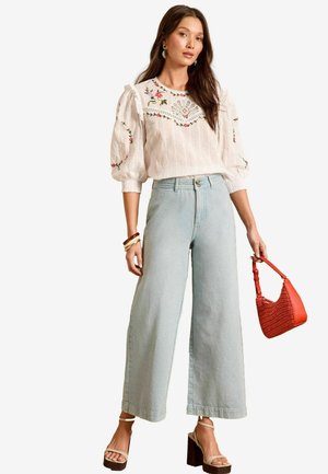 Blusa bianca ricamata con maniche a sbuffo, jeans larghi di un blu chiaro e una borsa intrecciata rossa. Sandali con tacco a cuneo completano l'outfit.