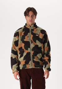 Giacca in pile con motivo mimetico verde, marrone e nero; presenta un colletto alto, chiusura con zip e tasche laterali. Materiale strutturato.