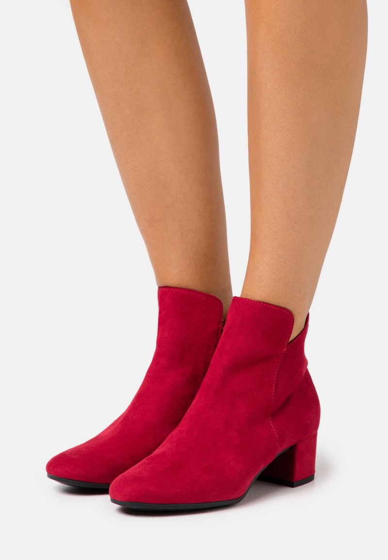 Paire de bottines en cuir suédé rouge à talons carrés portées sur des jambes nues sur un fond clair uni.