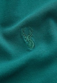 Tela verde azulado con un patrón texturizado y tejido que presenta un emblema de ciervo bordado en un verde ligeramente más oscuro.
