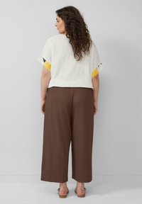 Witte top met korte mouwen en gele patroonaccenten, gecombineerd metbruine wijde broek. Model staat op een neutrale achtergrond.