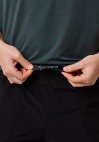 Donkergroene sportshirt met kleine perforaties, gecombineerd met zwarte shorts. Een trekkoord is gedeeltelijk uit de tailleband getrokken.