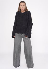 Schwarzer Kabelstrickpullover mit weiten Ärmeln, kombiniert mit hochtaillierten, weiten grauen Jeans, vor einem hellen Hintergrund.