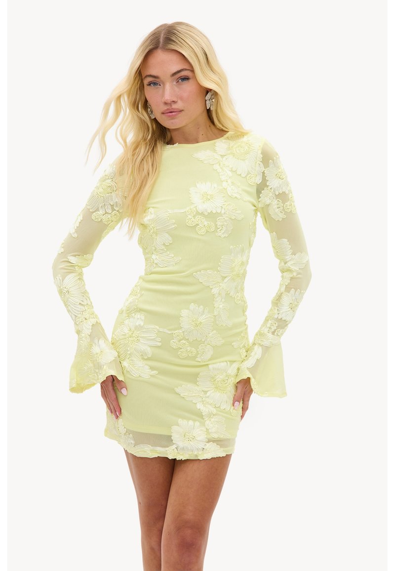 Femme blonde portant une robe jaune clair cintrée avec une broderie en dentelle florale et des manches longues évasées, debout avec les mains sur les hanches.