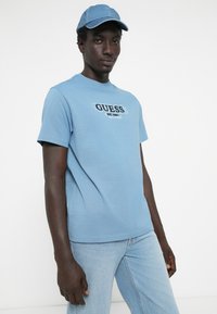 Modrá bavlnená tričko s nápisom "GUESS" v čiernej farbe. Doplní ju modrá džínová čiapka. Model má na sebe svetlomodré džínsy a stojí pred jednoduchým pozadím.
