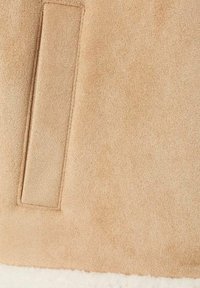 Beige Wildlederstoff mit einer weichen Textur und einer gesteppten Taschenapplikation, über einem weißen, flauschigen Futter entlang der unteren Kante geschichtet.
