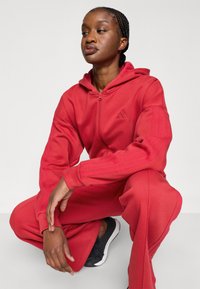 Červená mikina na zips a zodpovedajúce tepláky z mäkkej tkaniny. Má logo Adidas a tri pruhy na rukávoch. Čierne topánky dopĺňajú vzhľad.