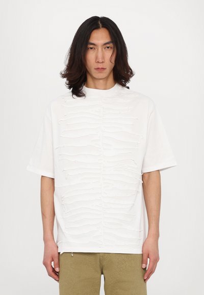 T-shirt bianco a maniche corte con linee orizzontali testurizzate sulla parte anteriore; scollatura rotonda; vestibilità comoda. Indossato con pantaloni beige.