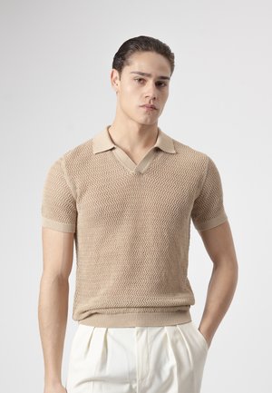 Joven con cabello peinado hacia atrás, vistiendo un polo de punto beige de manga corta y pantalones blancos plisados, de pie frente a un fondo liso.