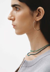 Pendiente de aro dorado usado por una persona con cabello oscuro, complementado con collares de cuentas turquesas en capas y un suéter de punto gris.