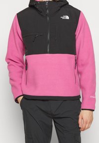 Chaqueta de forro polar con una sección superior negra y una sección inferior rosa. Presenta un bolsillo en el pecho con cremallera y bolsillos laterales, con capucha ajustable.