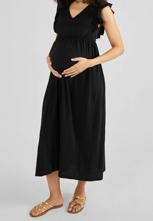 Gravid kvinde i sort ærmeløs kjole og guld flade sandaler, der holder sin mave mod en ensfarvet grå baggrund.