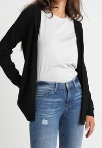 Black, open-front cardigan made of soft material, featuring long sleeves and a relaxed fit, worn over a white t-shirt and blue distressed jeans.
