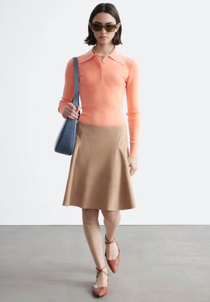 Kvinde går iført ferskenfarvet langærmet polo, tan A-formet nederdel, brune sko med ankelrem, solbriller, guldkædehalskæde og blå skuldertaske.