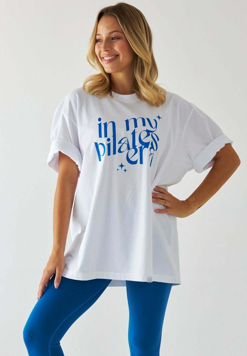 Biała, oversize'owa koszulka z podwiniętymi rękawami, z niebieską typografią "w mojej erze pilatesu". Połączona z niebieskimi legginsami, o gładkiej fakturze materiału.