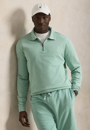 Άνδρας που φοράει γαλάζιο-πράσινο φούτερ με φερμουάρ στο 1/4 και αντίστοιχο παντελόνι φόρμας, λευκό καπέλο, στέκεται μπροστά σε μπεζ τοίχο με το ένα χέρι στην τσέπη.