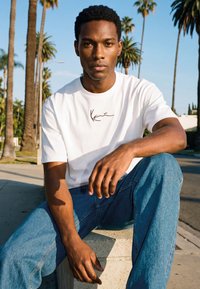 Joven con camiseta blanca y vaqueros azules sentado al aire libre con palmeras y un cielo despejado de fondo.