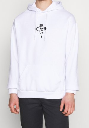 Persona con sudadera blanca con capucha, con caracteres japoneses negros y flechas en el pecho, combinada con pantalones negros, de pie frente a un fondo sencillo.