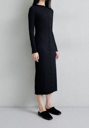 Femme portant une robe midi noire à manches longues avec des bords volantés et des chaussures noires faciles à enfiler, debout contre un mur clair uni.