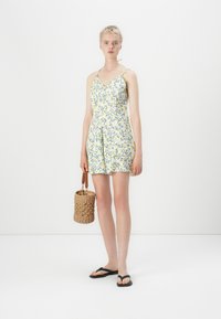 Blommönstrad jumpsuit i vitt, gult och grönt. Har spaghetti-remmar och cinchad midja. Kommer med en vävd väska och flip-flops.