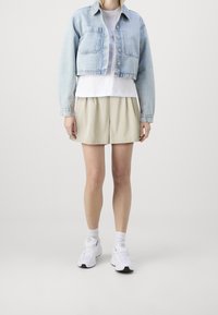 Veste courte en denim bleu clair, t-shirt blanc superposé dessous, short plissé beige et chaussures de sport blanches avec des accents noirs.