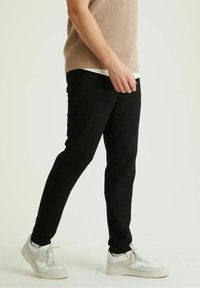 Muž v černých slim-fit kalhotách, bílých teniskách a béžové bezrukávové pletené vestě, stojící na pozadí s jednoduchým světlým tónem.