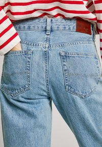 Light blue denim jeans feature a high waist, patch back pockets, and subtle fading. The waistband has a brown leather label detail.
